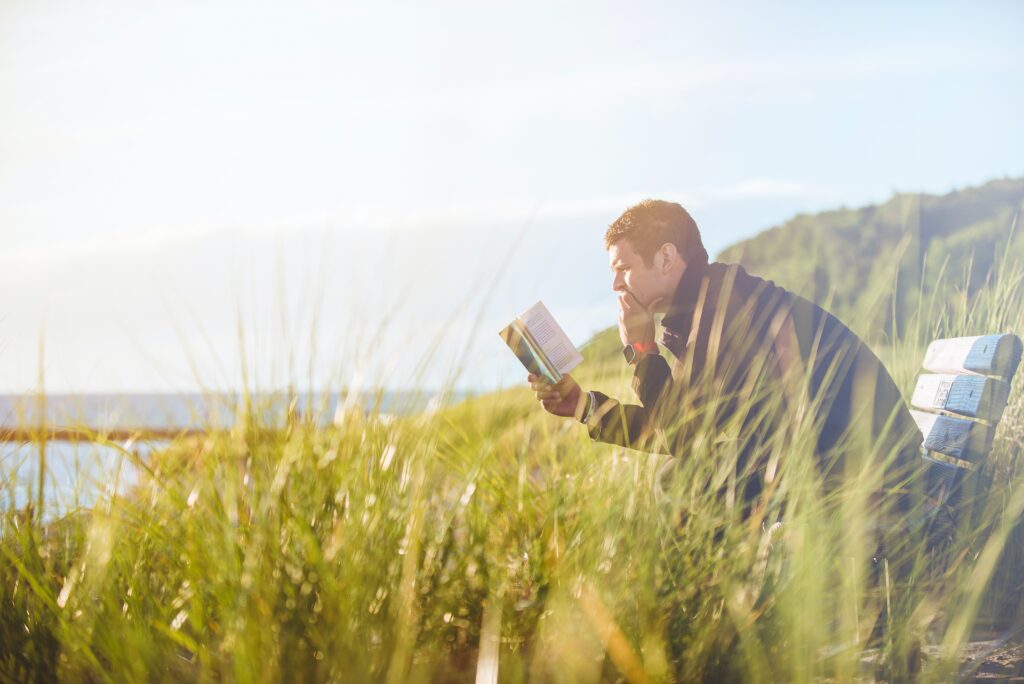 Hombre leyendo un libro sentado en una banca en medio de mucha vegetación.