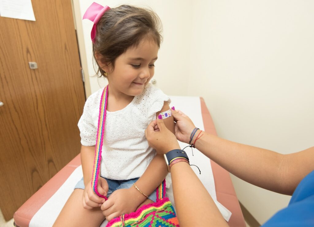 Foto de una niña que mira con una sonrisa, mientras una enfermera le pone un parche curita en su brazo izquierdo.