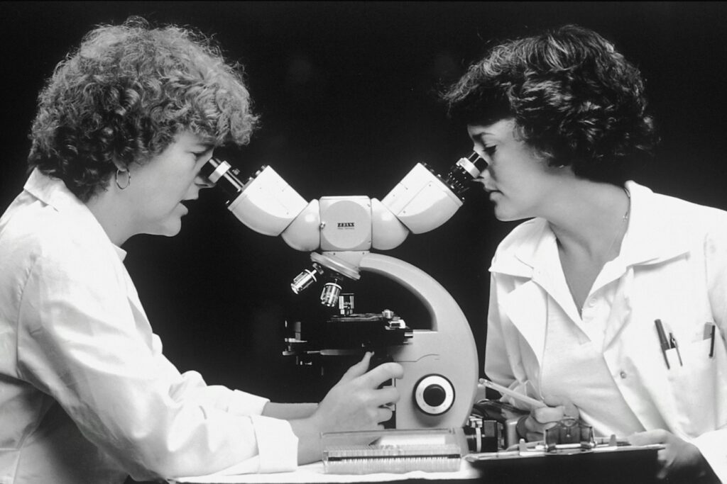 Foto en blanco y negro de dos mujeres científicas, de décadas pasadas, que miran por un microscopio.