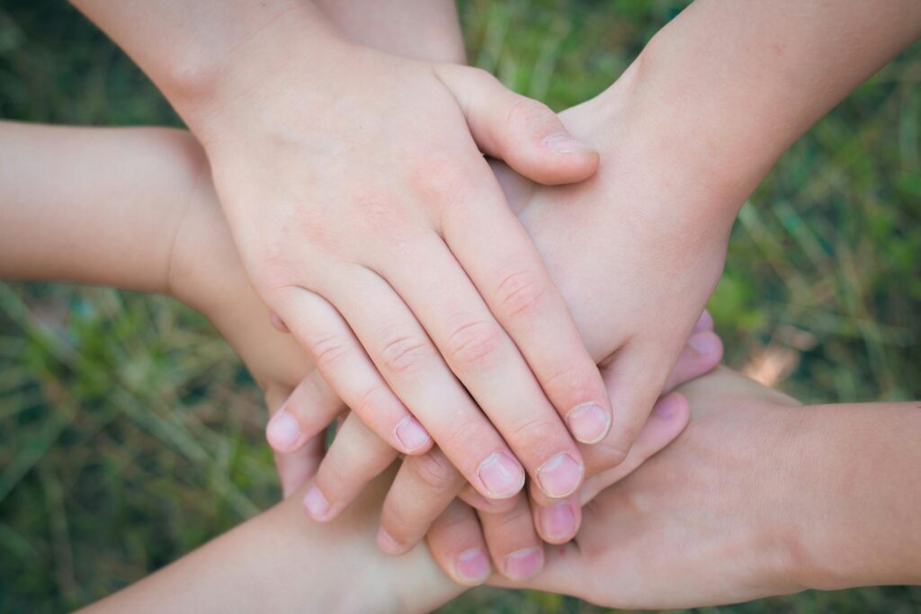 Foto que en el centro muestra varias manos unidas, unas sobre las otras, y en el fondo se ve pasto.