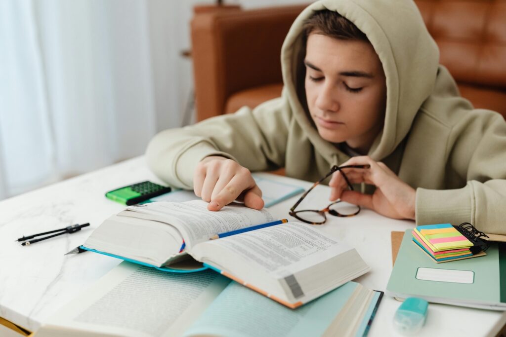 Foto de un joven estudiando sentado frente a un escritorio, donde hay libros abiertos, notas adhesivas, lápices y otros. El joven apunta el libro con su mano derecha y con la izquierda sostiene unos lentes.