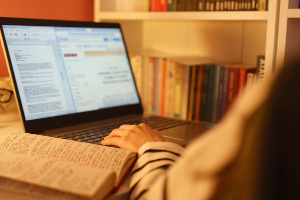 Foto de una persona que está trabajando en una laptop. En el lado izquierdo de la imagen se ve la laptop y un cuaderno abierto con apuntes, al lado derecho, solo el brazo izquierdo de la persona y en el fondo de la imagen un librero, lleno de libros.